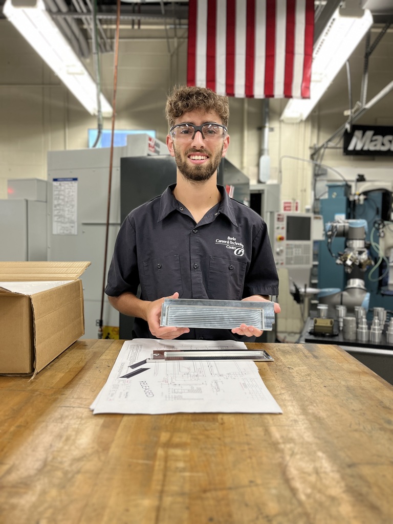A Berks Career & Technology Center Computerized Machining Technology student stands in a machining lab holding a finished metal satellite deployer door in one hand and a technical drawing in the other. An American flag hangs overhead, and a CNC machine is visible behind them.