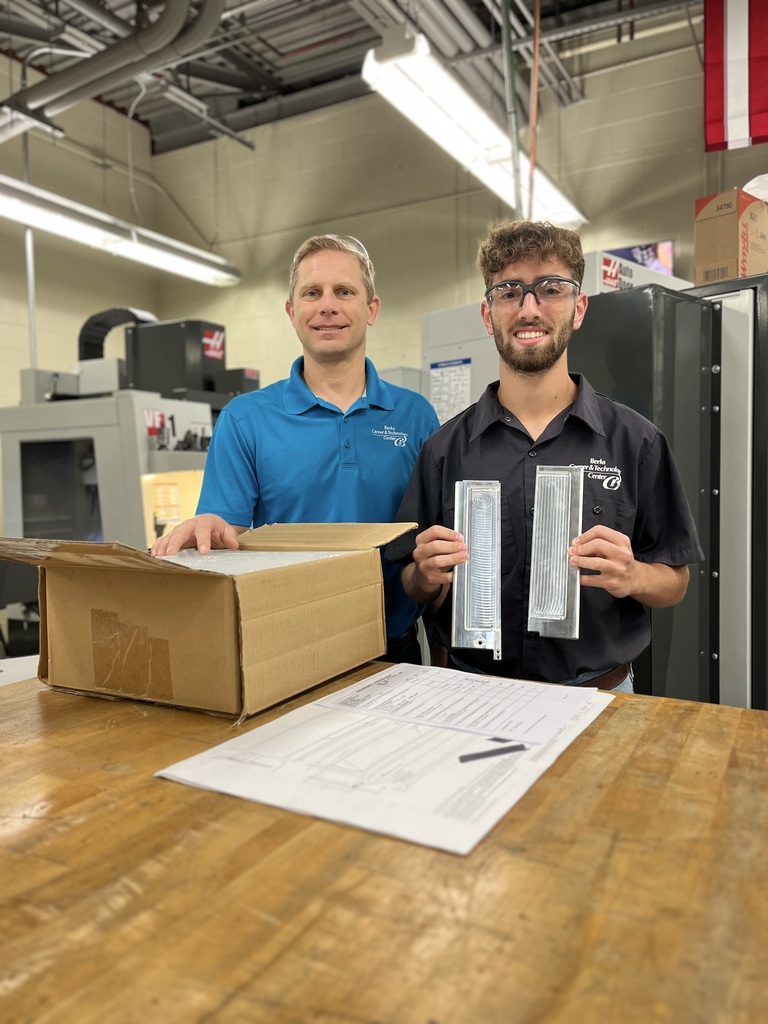 A Computerized Machining Technology student and instructor stand together in the machining lab at Berks Career & Technology Center. The student holds two finished metal satellite deployer doors, while the instructor stands beside an open box on a workbench. Technical drawings and machining tools are visible on the table.