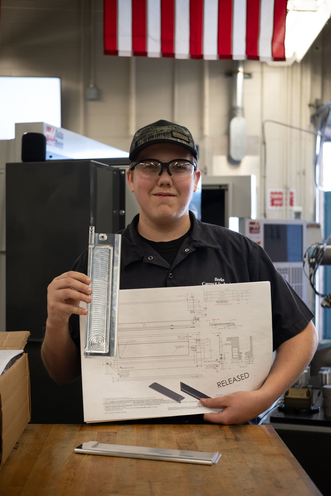 A Berks Career & Technology Center Computerized Machining Technology student stands in the lab holding a stack of finished metal satellite deployer doors. A technical drawing and open cardboard box sit on the workbench in front of him, with an American flag hanging above and machining equipment in the background.