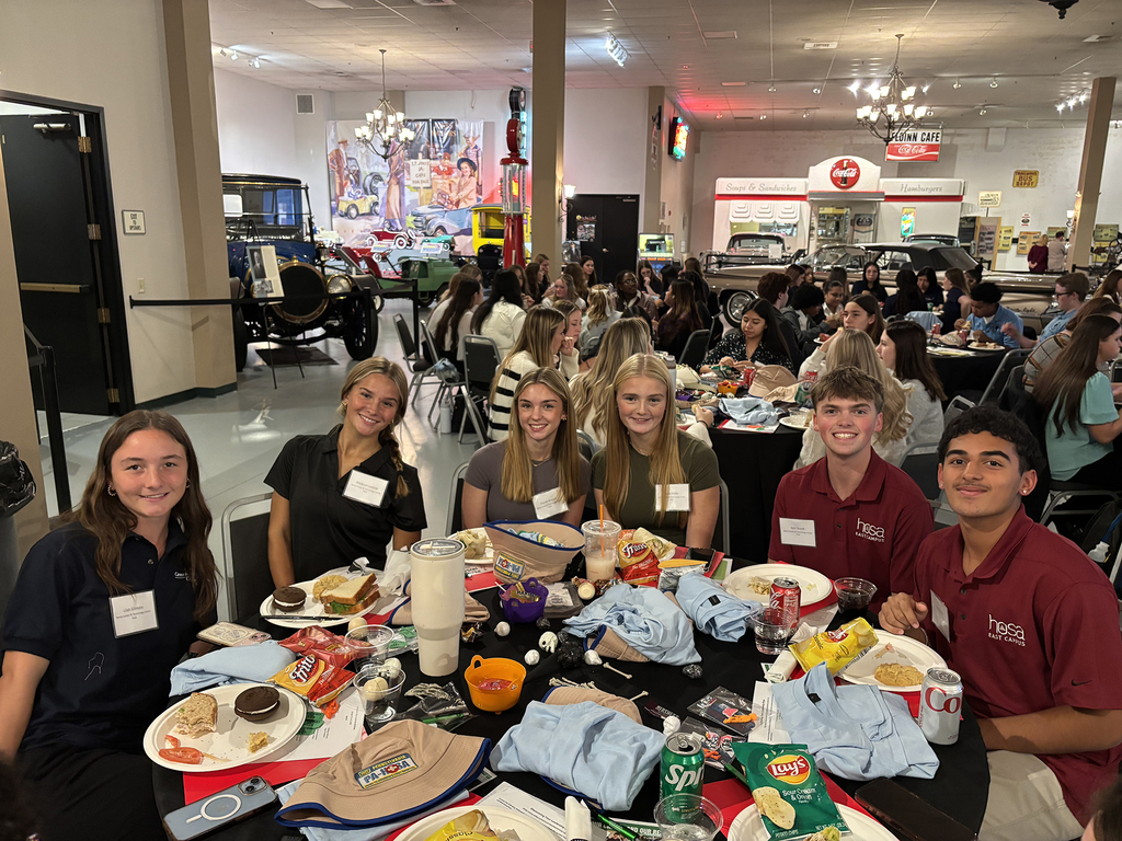 Six BCTC students gather around a lunch table inside a large room decorated with antique cars and retro signage. They’re enjoying food and drinks, wearing their name badges, and smiling for the photo.