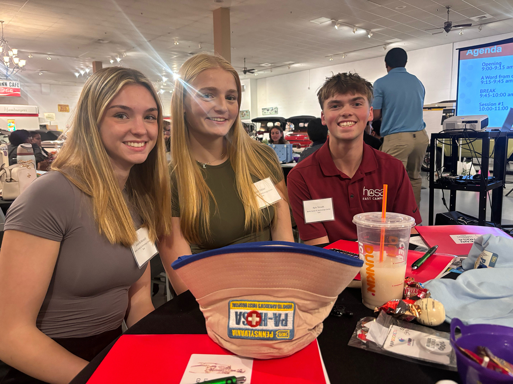 Three BCTC students pose for a photo at their table, smiling with conference materials, snacks, and a Dunkin’ drink visible in front of them. The event agenda is projected on a screen in the background.