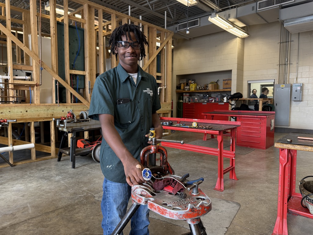 A first-year Plumbing and Heating student at BCTC wearing safety glasses and a dark green uniform shirt stands in the shop holding a pipe cutter at a red workbench. Framed wall structures and plumbing materials are visible in the background.