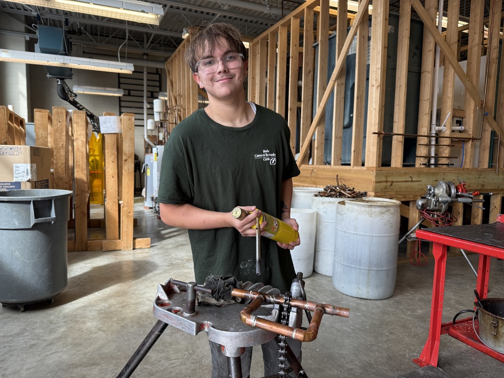 A BCTC Plumbing and Heating student stands at a red workbench holding a yellow torch and copper pipe assembly. Wooden framing and plumbing setups are in the background of the shop.
