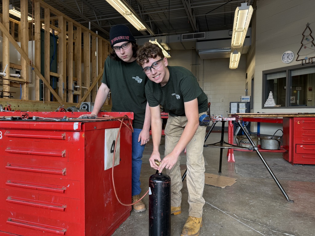 Two Plumbing and Heating students work together in the shop, standing near a red tool cabinet and handling a black gas cylinder used for soldering practice. Both are wearing safety glasses and green BCTC shirts.