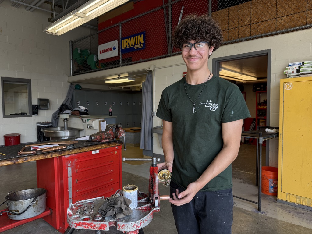 A student in the Plumbing and Heating program smiles while holding a small can of flux at a red workbench. He’s wearing safety glasses and a green BCTC shirt, with shop tools and lockers in the background.