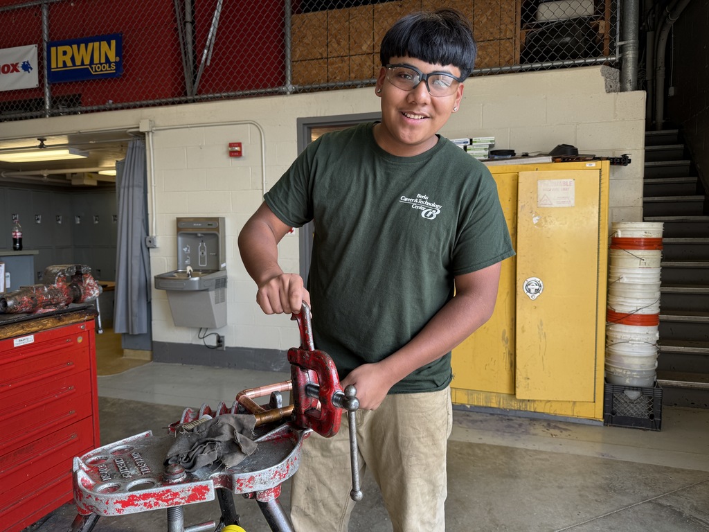 A student in BCTC’s Plumbing and Heating program smiles while operating a pipe vise in the shop. He’s wearing safety glasses and a green BCTC shirt, with yellow storage lockers and tool signs visible behind him.