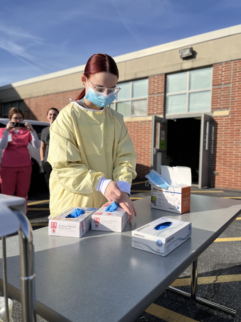 A Health Occupations student wearing a yellow isolation gown, mask, and safety goggles prepares to put on blue nitrile gloves at a table outside BCTC. Boxes of gloves and masks are laid out on the table. Two classmates in scrubs stand in the background smiling.