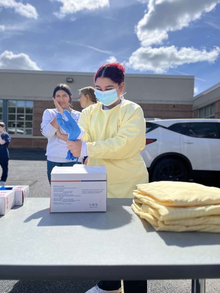 A Health Occupations student wearing a yellow gown, mask, and blue gloves adjusts one glove at an outdoor table. Boxes of protective gloves and gowns are on the table, while classmates look on in the background.