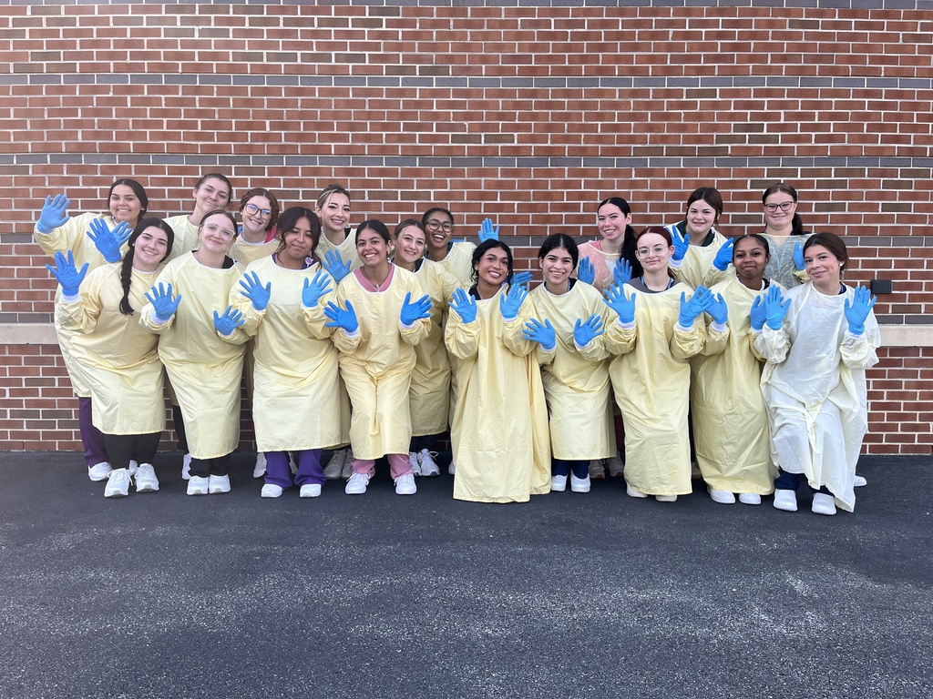 A large group of Health Occupations students stand together in front of a brick wall, all wearing yellow isolation gowns and blue gloves. The group smiles and waves toward the camera, showing teamwork and enthusiasm.