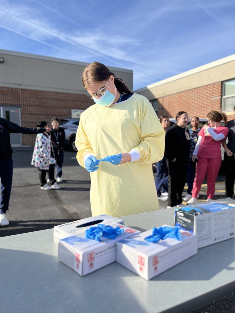 A Health Occupations student dressed in a yellow isolation gown and blue gloves focuses on donning gloves at an outdoor table. Several classmates and instructors stand behind her, watching and smiling, with the school building visible in the background.