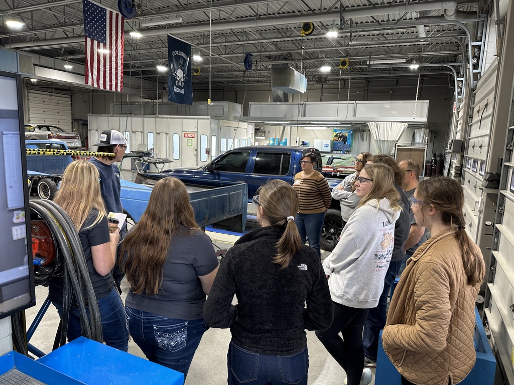 A group of visitors stands around an instructor and students in the Collision Repair Technology lab. A blue pickup truck and large equipment are visible in the background under hanging American flags.