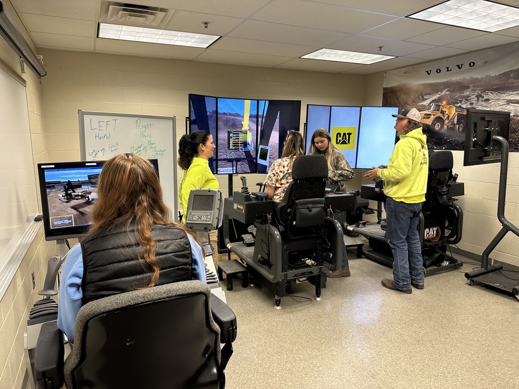 Visitors observe the Heavy Equipment Operations classroom, where simulators display virtual construction equipment. A student sits at a CAT simulator while others look on.
