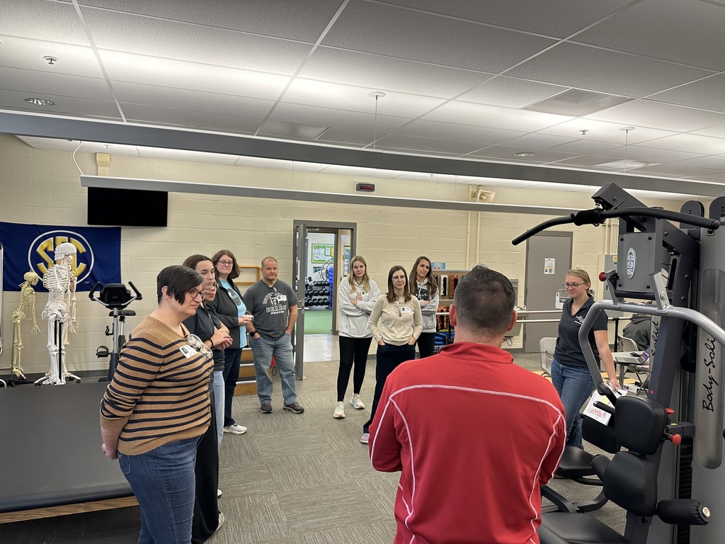 A BCTC instructor speaks to a group of visiting educators inside the Exercise Science & Rehabilitation program. Gym and therapy equipment, including a weight machine and anatomical skeleton, are visible in the background.