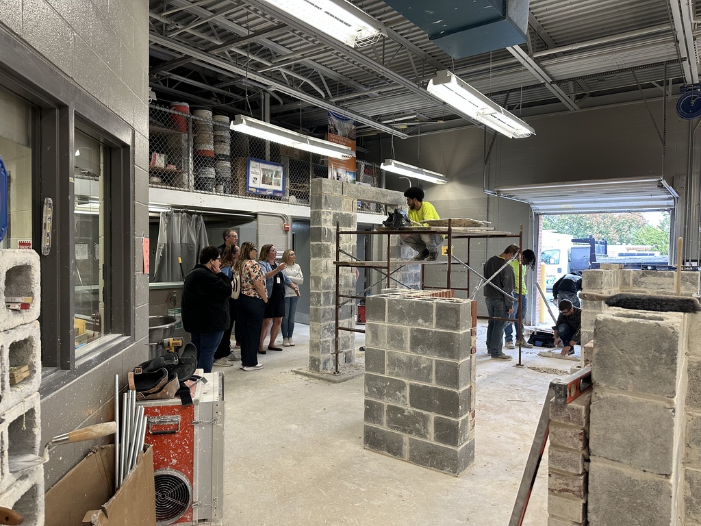 Educators watch BCTC Masonry students actively working on block and brick structures inside the masonry lab. The space includes workstations, scaffolding, and partially completed walls.
