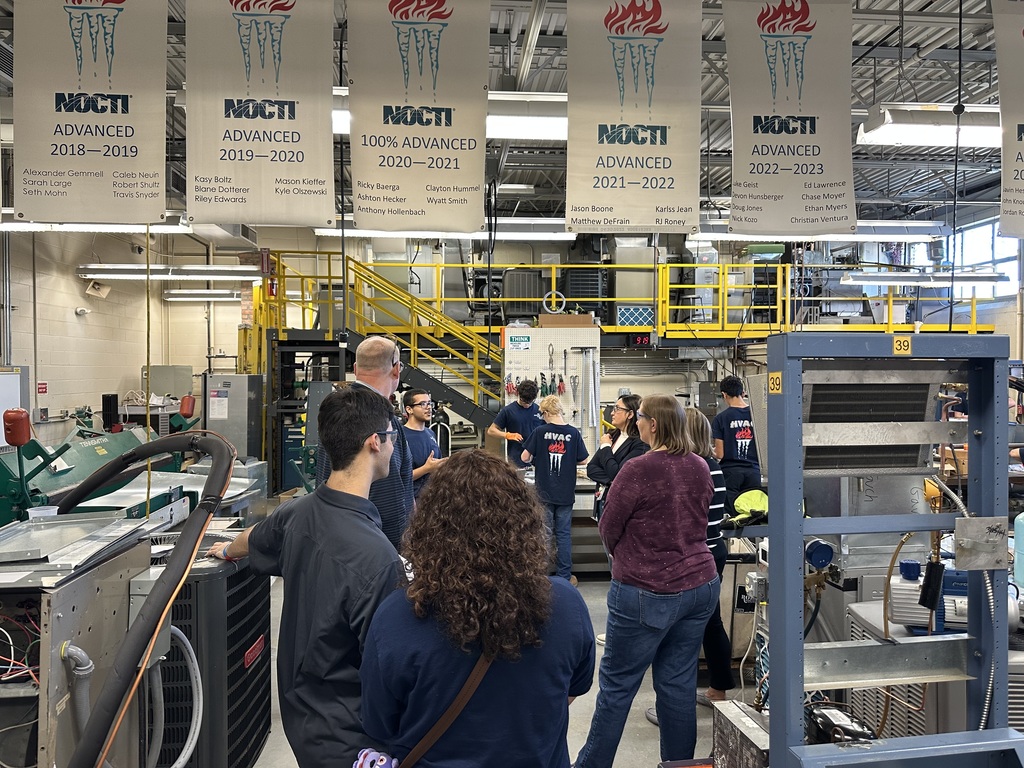 A group of visitors stands inside BCTC’s HVAC program lab, listening to a student or instructor. HVAC equipment, ductwork, and NOCTI Advanced banners hang above the workspace.