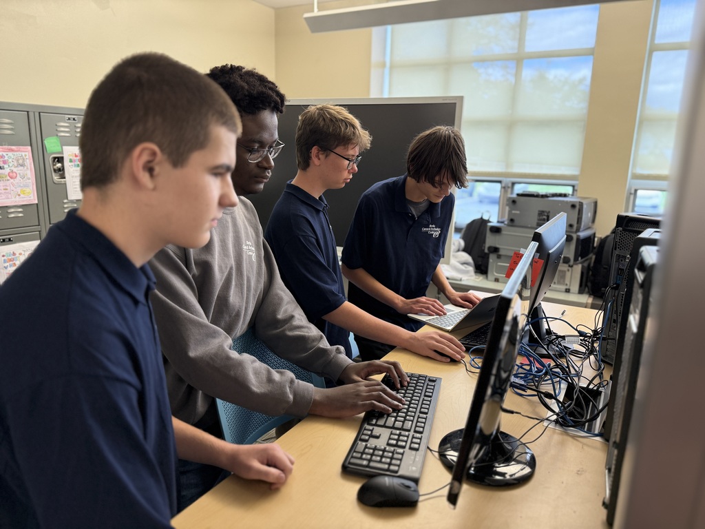 Four Computer Systems Networking & Security students work together at desktop computers, typing on keyboards and looking toward the camera in a bright classroom with large windows.