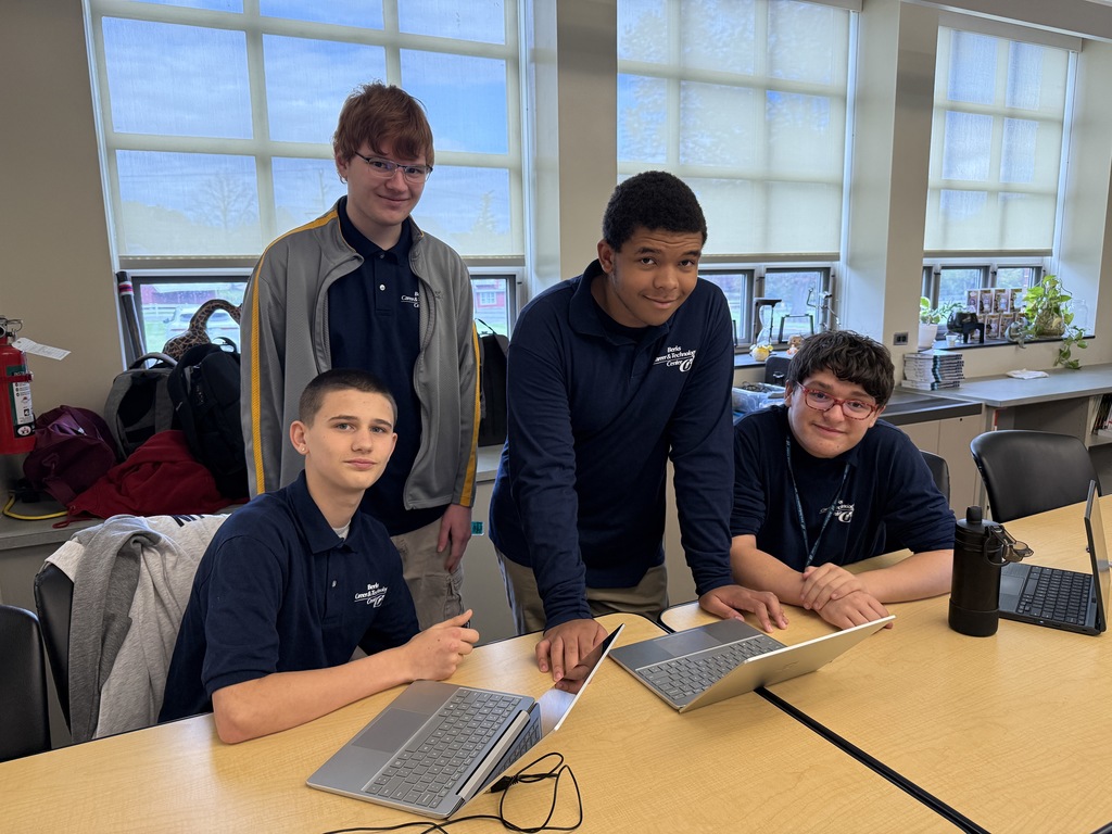 Four Computer Systems Networking & Security students sit and stand around a classroom table with laptops open, smiling and looking at the camera in front of bright windows.