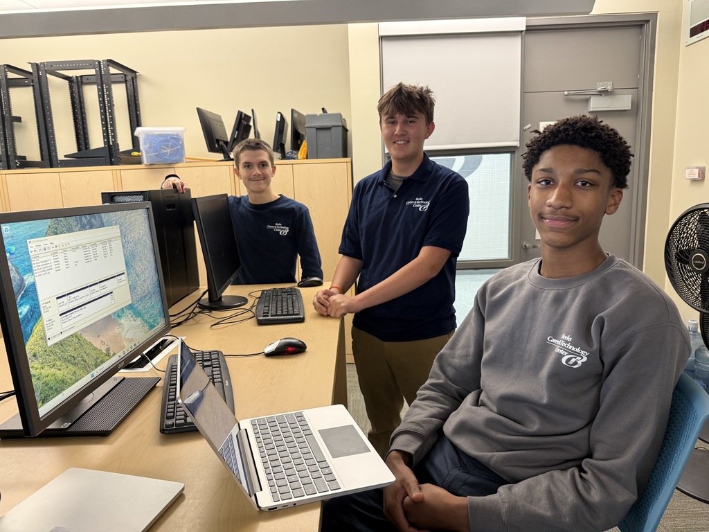 Three Computer Systems Networking & Security students pose at their workstation in a classroom, with computer monitors, a laptop, and networking equipment on the desk in front of them.