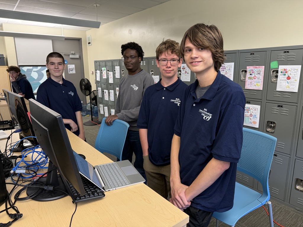 Four Computer Systems Networking & Security students at Berks Career & Technology Center stand beside computer monitors and a laptop in their classroom. They are smiling at the camera while surrounded by lockers and network cables on the table.