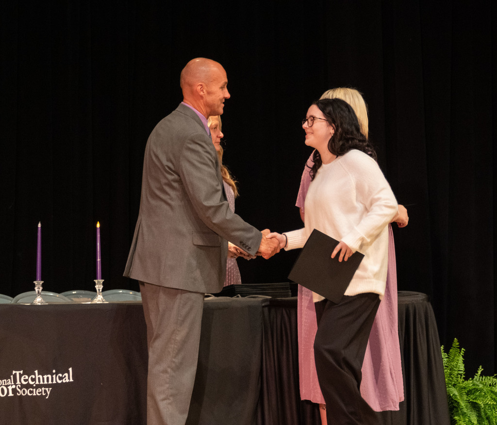 A female student wearing glasses and a white sweater shakes hands with Dr. Stauffer on stage while receiving her National Technical Honor Society certificate during the induction ceremony.
