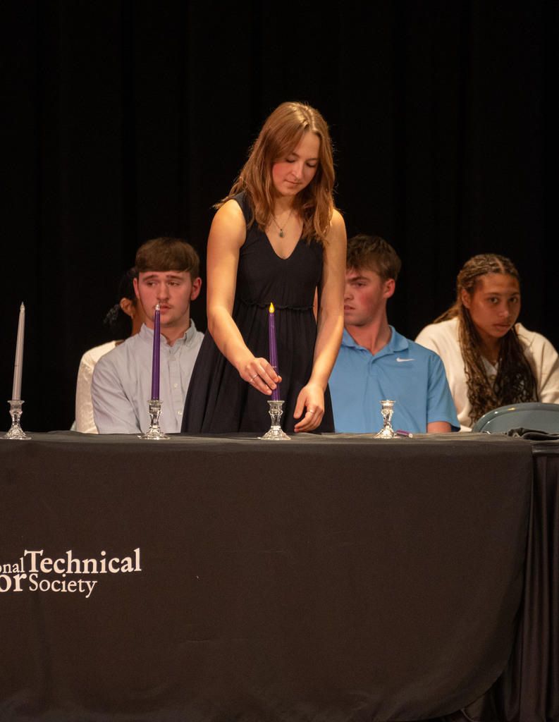 A student in a black dress lights a candle on stage as part of the National Technical Honor Society Induction Ceremony while other inductees watch from behind the table draped with the NTHS logo.
