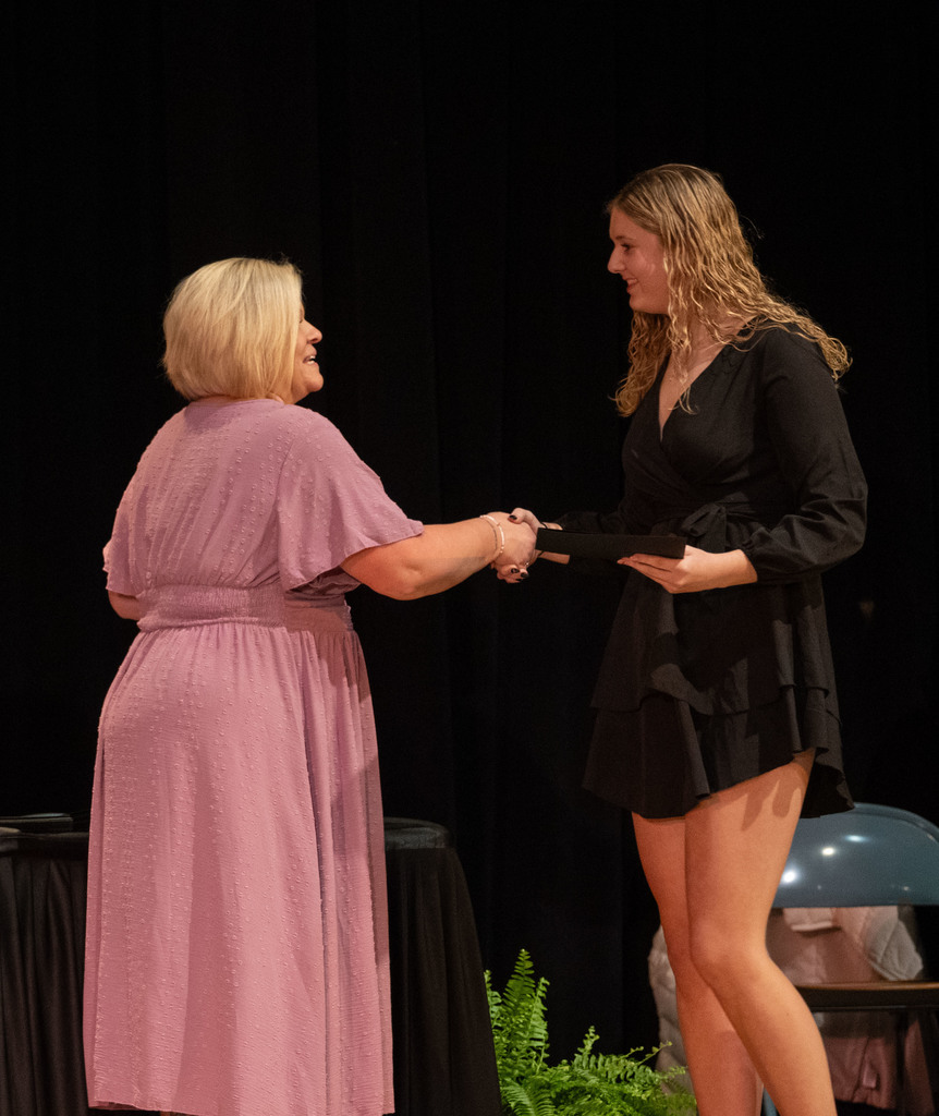 A Berks Career & Technology Center staff member in a lavender dress congratulates a student in a black dress on stage during the National Technical Honor Society Induction Ceremony.