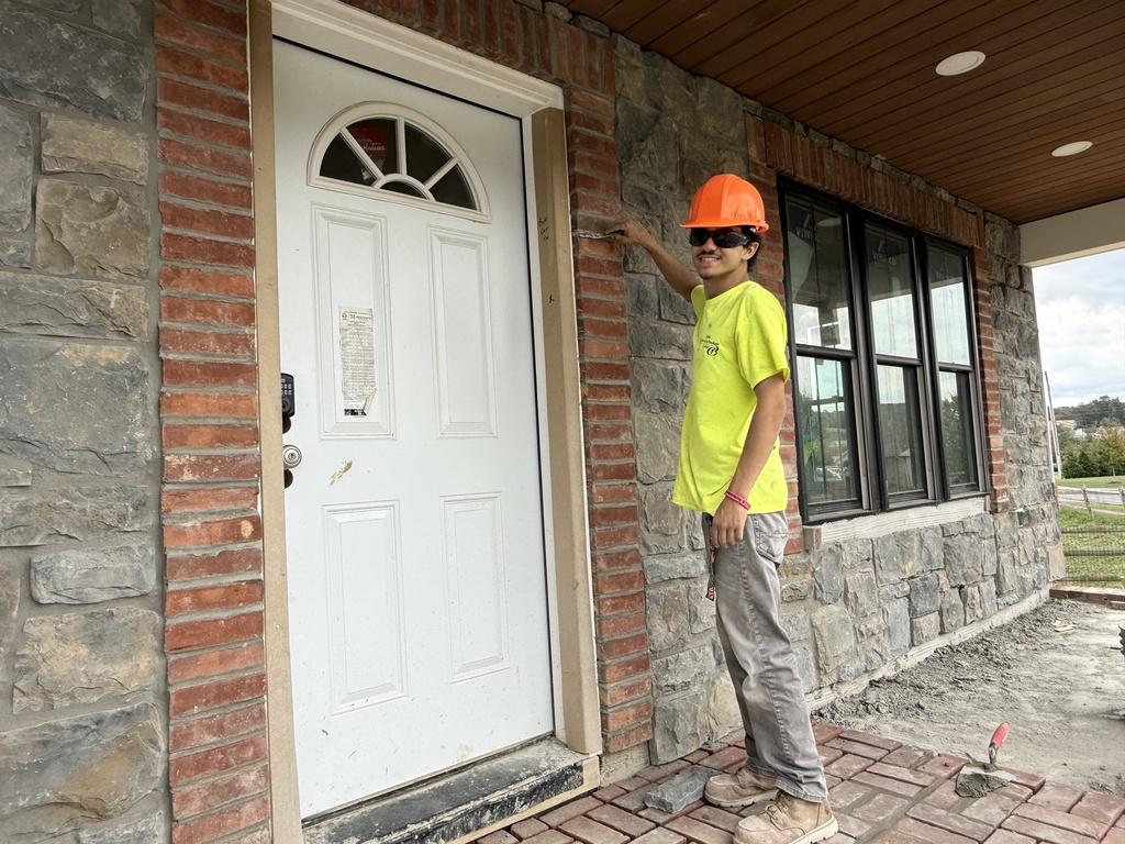 A Masonry student wearing a yellow safety shirt, orange hard hat, and safety glasses stands beside a white door, using a masonry tool on the wall’s red brick trim. The stone veneer wall and brick porch beneath him are part of the Student Built House.