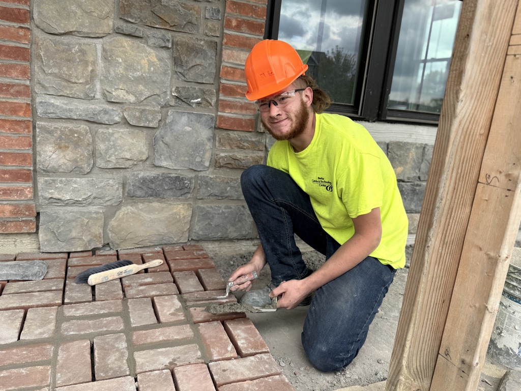 A Masonry student in a yellow safety shirt and orange hard hat crouches on the brick porch while filling mortar joints between bricks. The stone veneer wall and red brick sill are visible behind him, along with masonry tools on the ground.