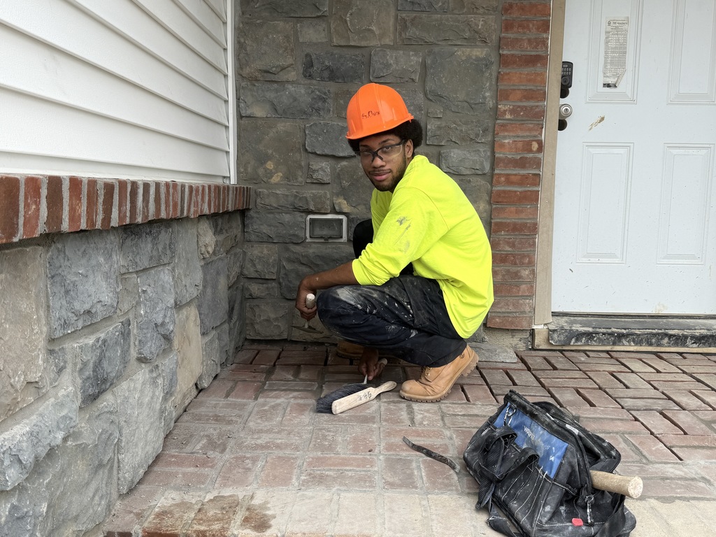 A Masonry student wearing a bright yellow safety shirt, orange hard hat, and safety glasses kneels on a newly laid brick porch. He’s holding a masonry tool and working near a stone veneer wall with a red brick sill above it.
