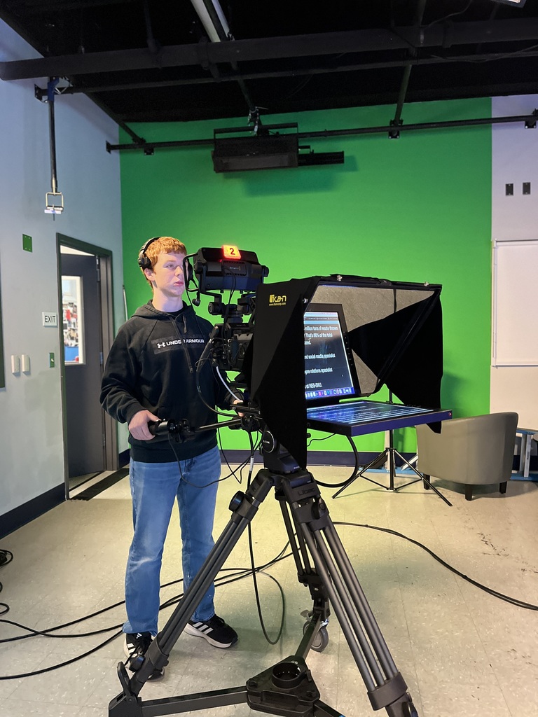 A student adjusts a studio camera and teleprompter setup in front of a green screen, wearing a headset and reading from the teleprompter screen.