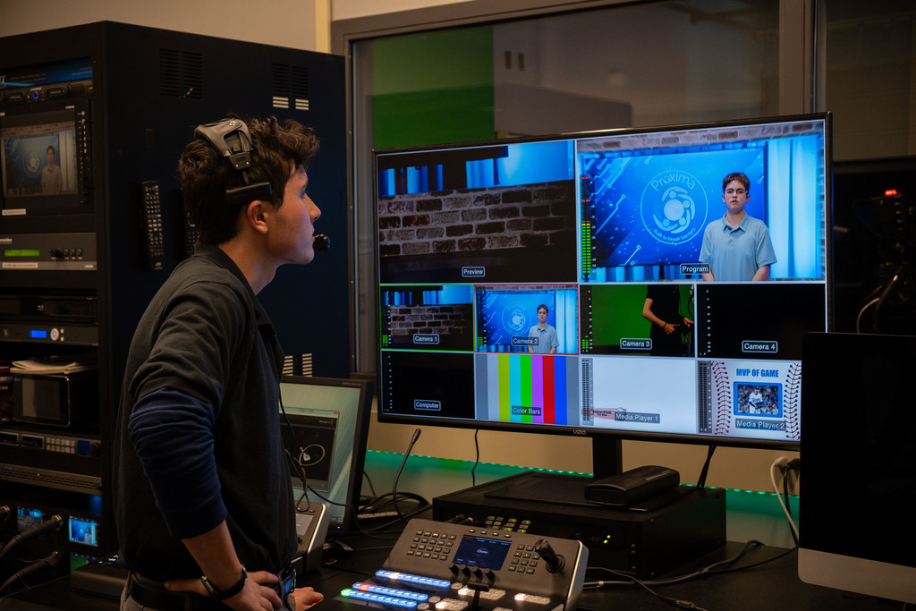 A student in a control room watches multiple live camera feeds on monitors while operating a video switcher and wearing a headset.