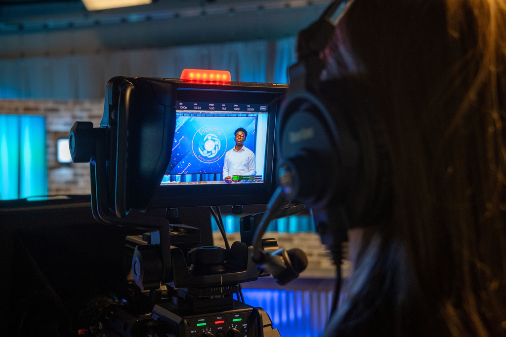 A student wearing headphones films another student who is presenting on a studio set in front of a “Proxima” logo displayed on a blue digital background.
