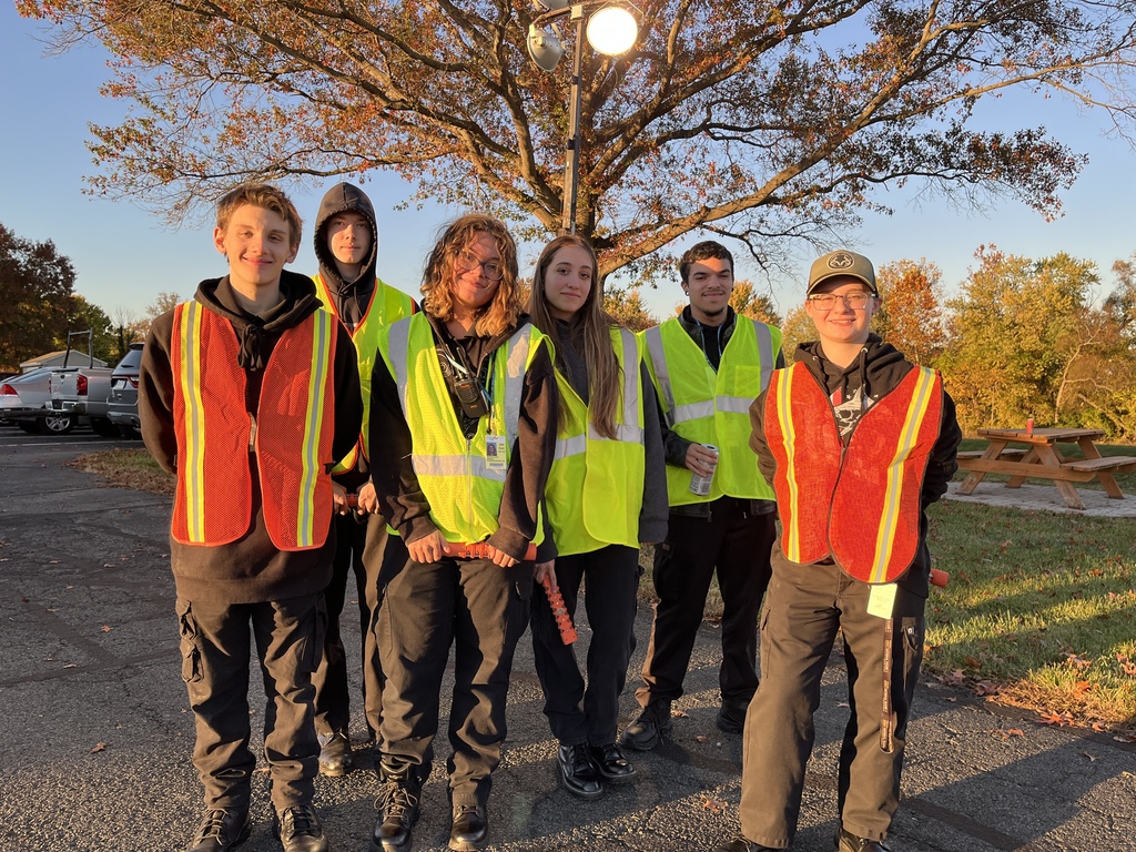 A group of seven Protective Services students wearing reflective vests pose outdoors at sunset. They are standing near a tree with fall foliage, with a picnic table and grassy area behind them.
