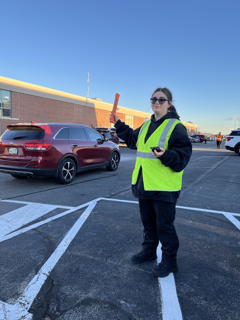A Protective Services student wearing a yellow reflective vest and sunglasses directs vehicles in a parking lot, holding an orange traffic baton. A red SUV is driving past, and the school building is visible behind her.