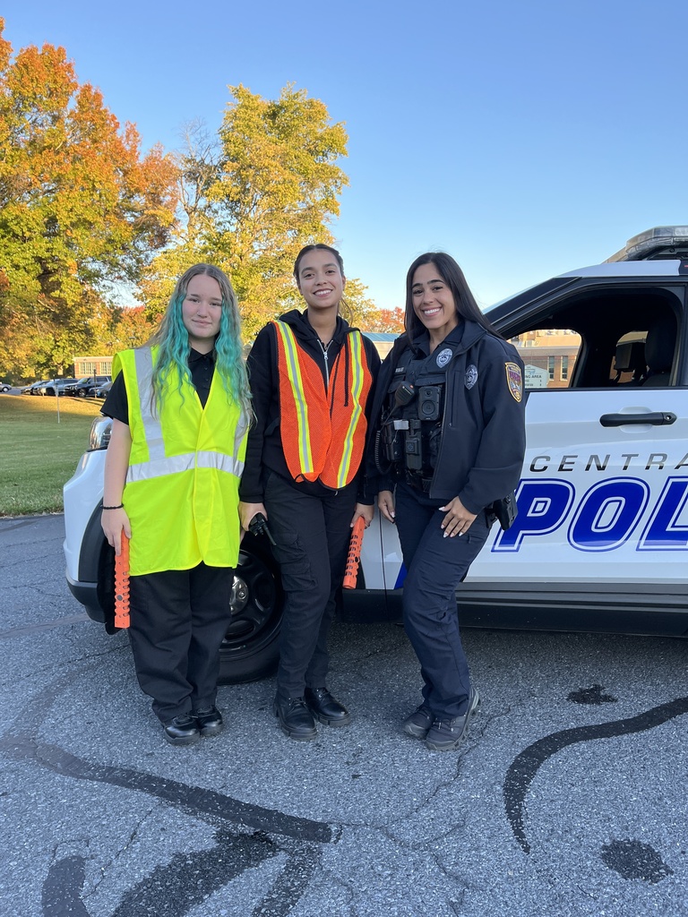 Two Protective Services students in reflective vests stand smiling next to a police officer in uniform. They are standing in front of a marked police SUV with autumn trees in the background.
