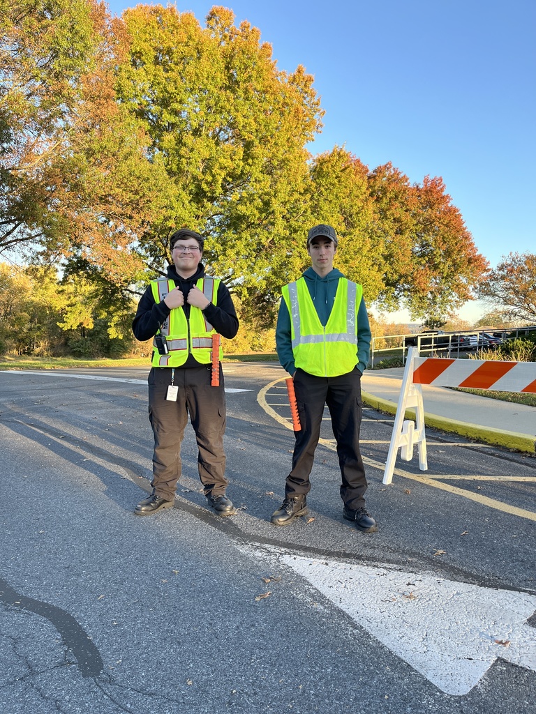 Two Protective Services students stand on a driveway in bright yellow reflective safety vests, holding orange traffic batons. Behind them are large trees with colorful fall leaves and a clear blue sky.