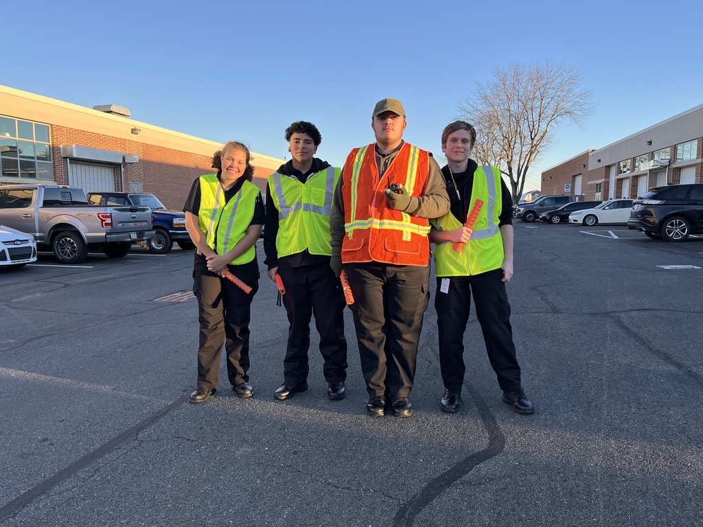 Four Protective Services students stand together in a parking lot, wearing reflective vests and holding orange traffic batons. The school building and parked cars are visible behind them in evening sunlight.
