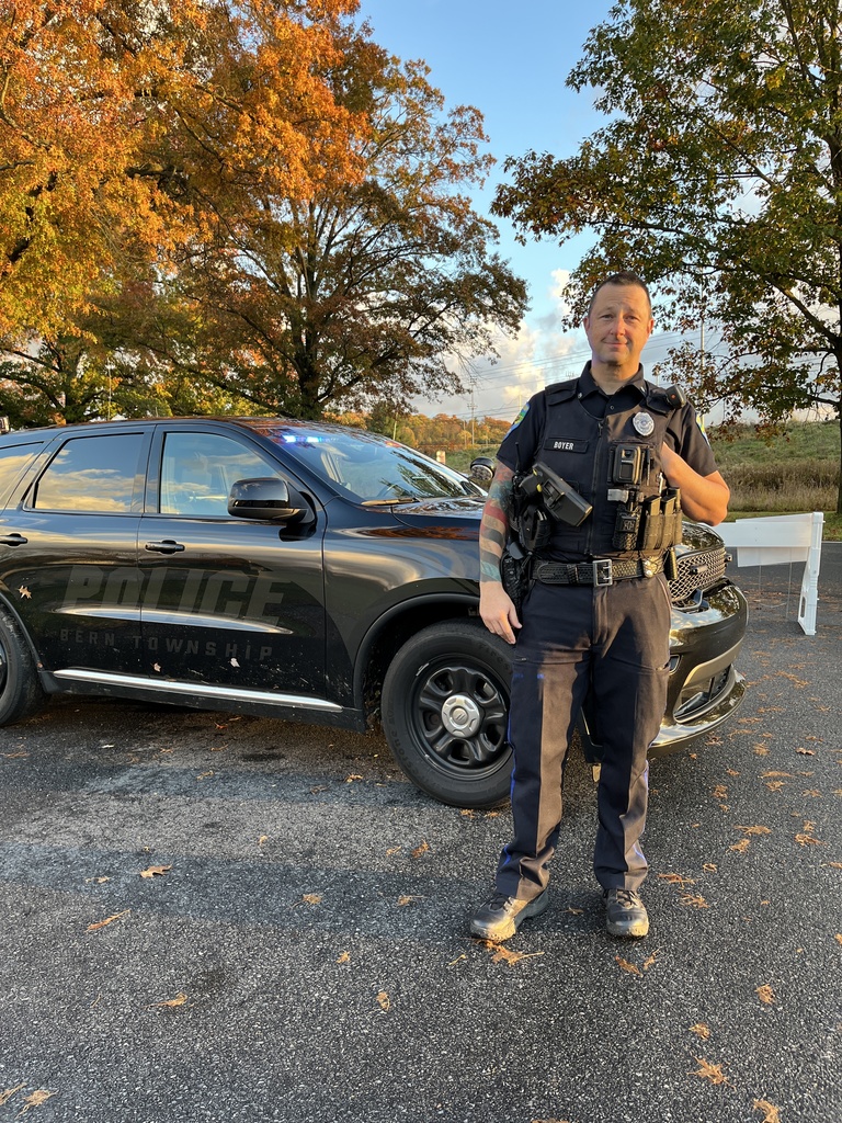 A police officer stands beside a marked Bern Township Police SUV in a parking lot during sunset. He is wearing a police uniform and equipment, with autumn trees in the background.