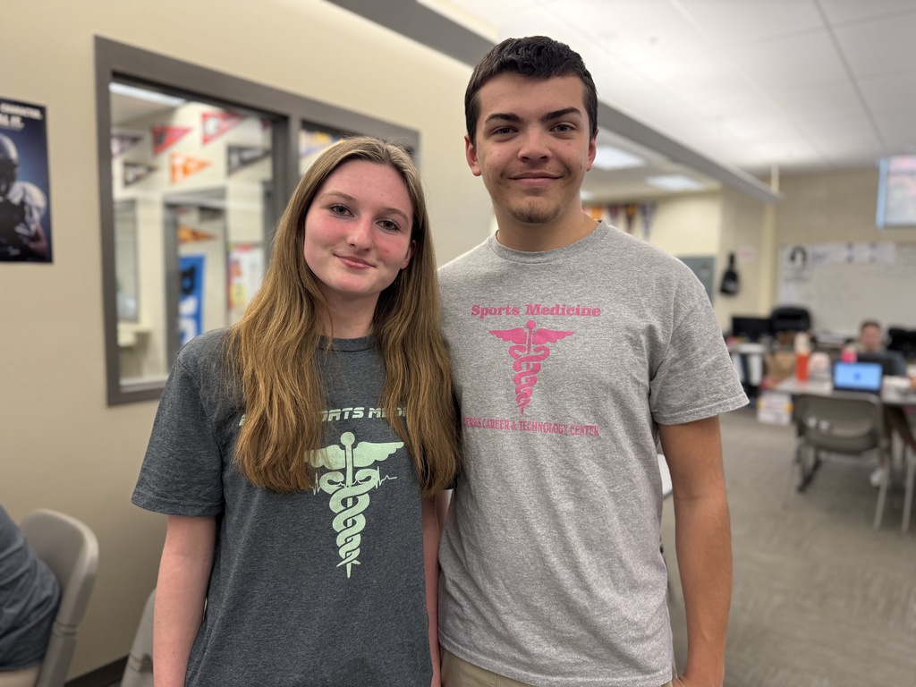 Two BCTC students stand side by side in a classroom, smiling at the camera. Both are wearing T-shirts with a medical caduceus graphic representing the Sports Medicine program. The classroom background includes desks, chairs, posters, and other students working.