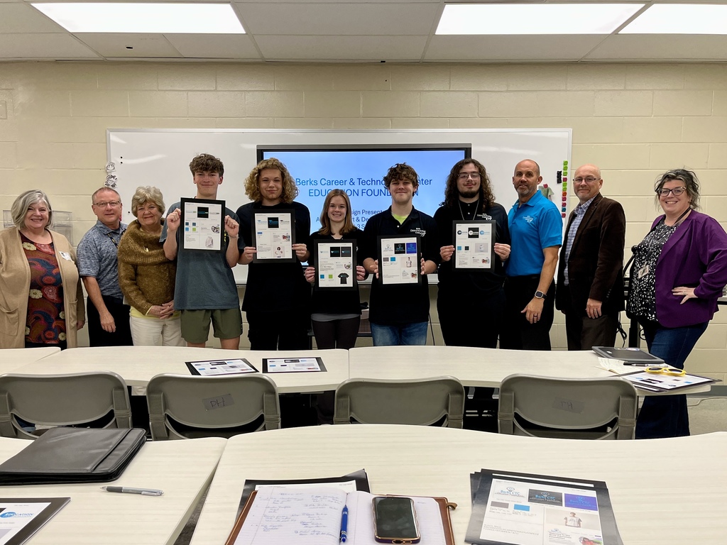 A group photo of students holding printed logo design boards, standing with instructors and school leaders in a classroom. A screen in the background displays “Berks Career & Technology Center Education Foundation Logo Design Presentations.” Tables and student design boards are visible in the foreground.