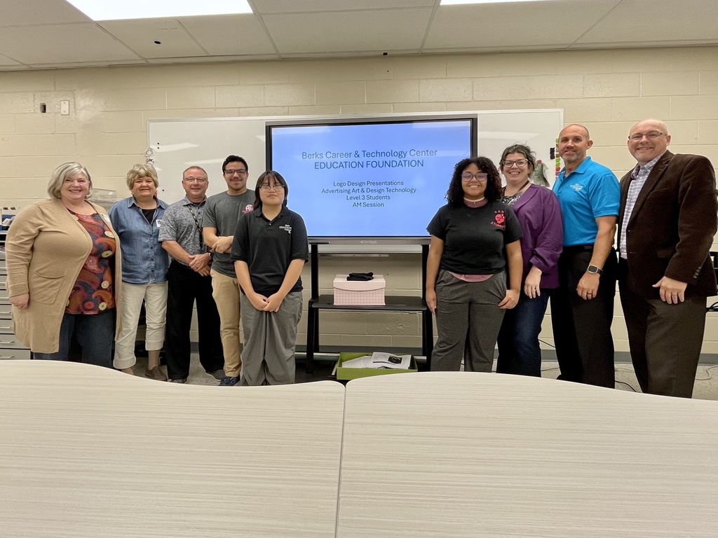 A group photo of students, instructors, and school representatives standing in a classroom in front of a screen displaying “Berks Career & Technology Center Education Foundation Logo Design Presentations.” Tables are visible in the foreground.