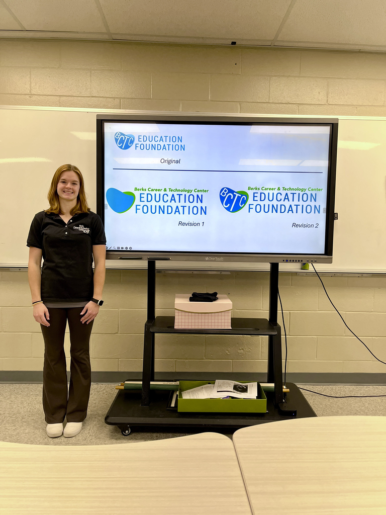 A student stands in a classroom smiling next to a large screen showing an original and two revised Berks CTC Education Foundation logos featuring heart shapes and circular badge-style designs. Tables are visible in the foreground.