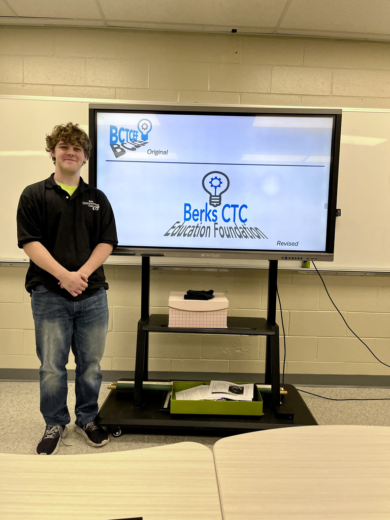 A student stands in front of a screen showing the original and revised Education Foundation logos featuring a lightbulb and gear concept. Tables are visible in the foreground.