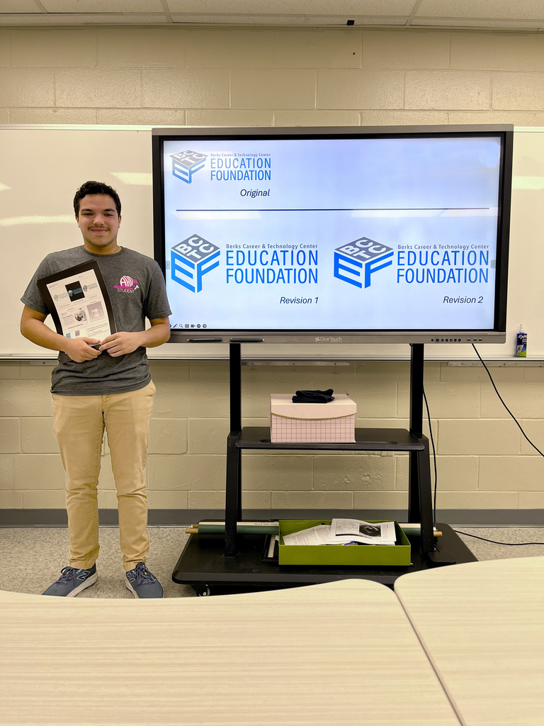 A student stands in a classroom holding a printed design sheet, smiling. Behind them is a large screen displaying three variations of a logo for the Berks Career & Technology Center Education Foundation labeled “Original,” “Revision 1,” and “Revision 2.” Tables are visible in the foreground.
