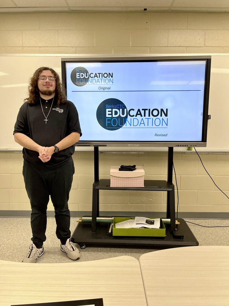 A student stands beside a screen showing the original and revised versions of a circular logo for the Berks CTC Education Foundation in blue and black tones. Tables are visible in the foreground.