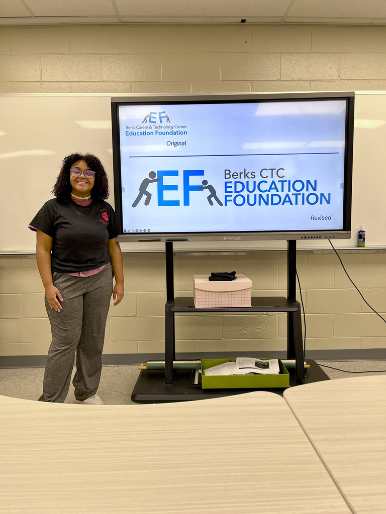 A student stands in a classroom and smiles beside a screen showing the original and revised versions of a Berks CTC Education Foundation logo featuring stylized figures pushing the letters “EF.” Classroom tables are visible in the foreground.