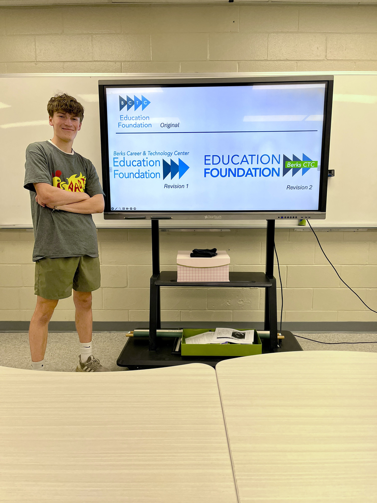 A student stands in front of a classroom screen displaying the original and revised Education Foundation logo concepts using arrow shapes and bold blue and green text. Tables are visible in the foreground.