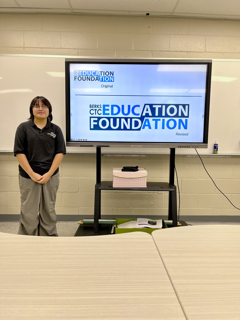 A student stands in front of a large screen showing the original and revised versions of a Berks CTC Education Foundation logo, with bold block text and a modern, blue color scheme. Tables are visible in the foreground.