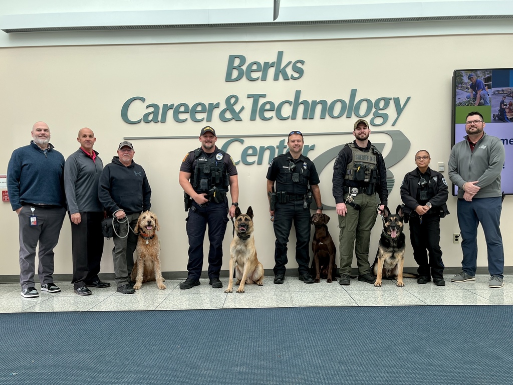 A group of nine adults stand in a line inside Berks Career & Technology Center, posing in front of a large wall sign with the school’s name. Several law enforcement officers are dressed in uniforms and safety gear, each accompanied by a canine partner, including a golden retriever, a Belgian Malinois, a chocolate Labrador, and a German Shepherd. Two school staff members stand at each end of the group. Everyone is facing the camera and smiling, with the dogs sitting calmly at their handlers’ sides.