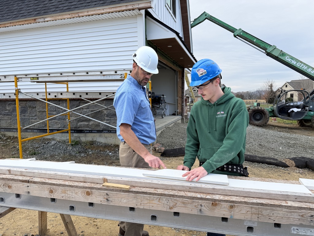 A Carpentry instructor wearing a white hard hat points to a frieze board on a workbench while a student in a blue hard hat and green hoodie listens and reviews the material. The Student Built House and scaffolding are behind them.